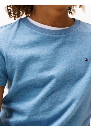 Dziecko o kręconych włosach, ubrane w białą podkoszulkę i jasno niebieski T-shirt z krótkim rękawem, z małym logo flagi na klatce piersiowej.