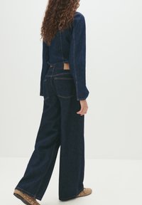 Woman with long curly hair wearing a dark denim jacket and wide-leg jeans, standing with one hand relaxed by her side.