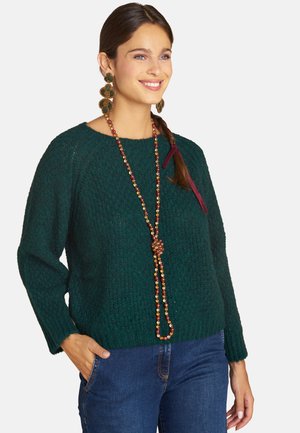 Frau lächelt, trägt einen dunkelgrünen gestrickten Pullover, eine lange Perlenkette, große Kleeblatt-Ohrringe und blaue Jeans mit einer Hand in der Tasche.