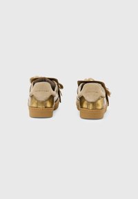 Un paio di sneakers beige e oro metallico con suole in gomma, viste da dietro su uno sfondo grigio chiaro e semplice.