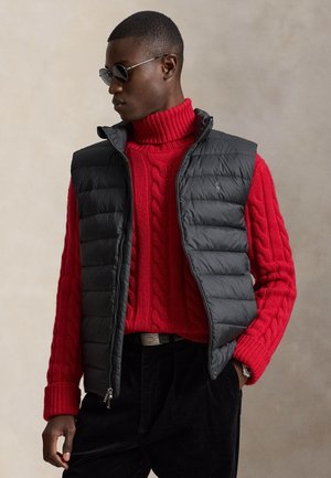 Homme portant des lunettes de soleil noires, un pull à col roulé à torsades rouge, un gilet matelassé noir et un pantalon noir, posant les mains dans les poches.