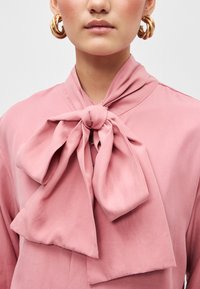 Blusa rosa feita de tecido suave, com um laço grande no colarinho, textura lisa e design minimalista sem padrões visíveis.