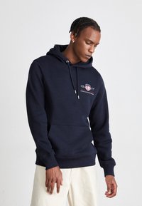 Marinblå hoodie med framficka, snörning i huvan och broderad logotyp. Tillverkad av mjukt, texturerat tyg för komfort.