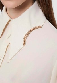 Blusa color crema con un colletto prominente e un accento curvo in tono oro impreziosito da strass chiari.