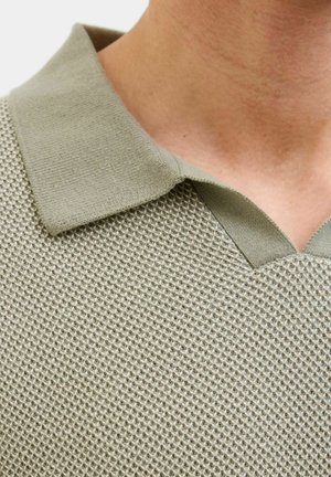 Tuva persona, kas valkā gaiši zaļu teksturētu trikotāžas polo kreklu ar ribotu apkakli un V veida izgriezumu.