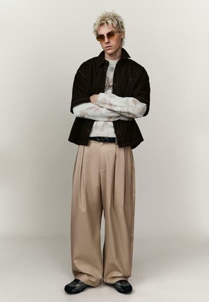 Osoba s blond vlasmi a slnečnými okuliarmi má na sebe čiernu bundu, biely sveter s vzorom, široké béžové nohavice a čierne tenisky, stojí so založenými rukami.