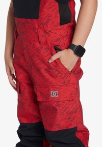Röda cargo-byxor med ett mönster av svarta klotter, solid svarta detaljer och en DC-logotyp. Inkluderar fickor och en hållbar tygtextur.