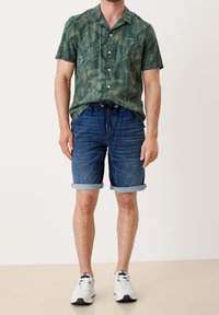 Hombre que lleva una camisa de manga corta verde con estampado de hojas, pantalones cortos de mezclilla enrollados y zapatillas blancas, de pie contra un fondo liso.