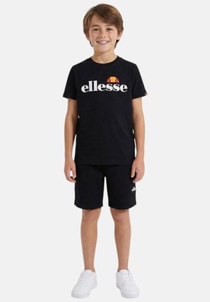 Jeune garçon debout, portant un t-shirt noir ellesse et un short assorti avec des baskets blanches, souriant sur un fond uni.