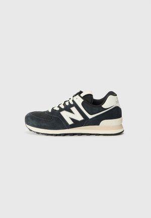 Sort-hvid New Balance sneaker med ruskindsoverdel, hvidt "N"-logo, hvide snørebånd og beige sål mod en ensfarvet lys baggrund.