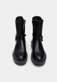 Botas de cuero negro hasta el tobillo con suela plana, cierre de cremallera lateral y correa decorativa con acentos metálicos. Textura suave en toda su superficie.