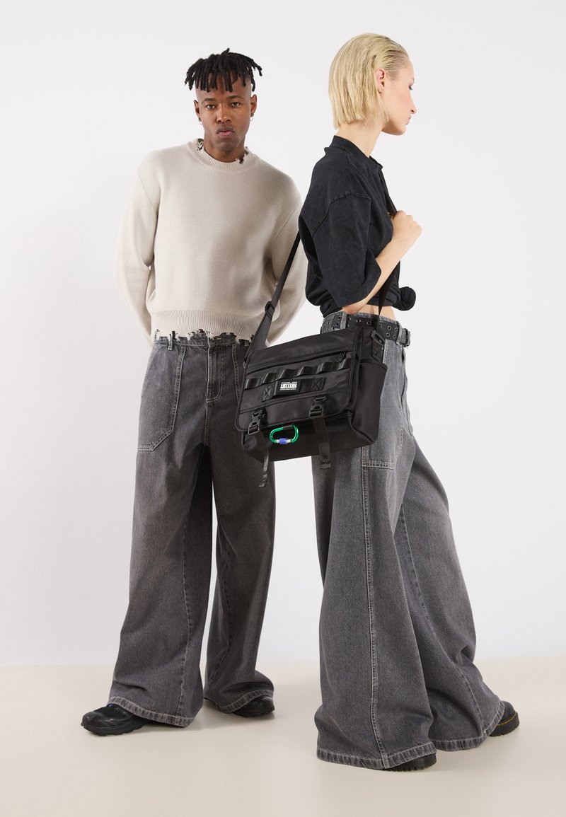 Bolsa mensajera negra con correa ajustable y tela texturizada, con múltiples bolsillos. Pareja de jeans grises de gran tamaño con dobladillo deshilachado.