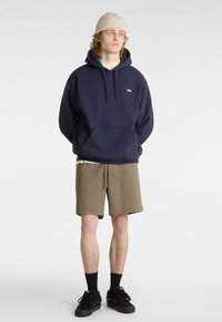 Marineblaue Kapuzenjacke mit Kängurutasche vorne und Kordelzügen, kombiniert mit olivfarbenen Shorts. Das Modell trägt schwarze Sneakers und eine beigefarbene Strickmütze.