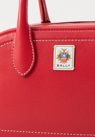 Sac à main en cuir rouge avec des coutures blanches, featuring un patch logo en or et émail. Poignées arrondies et forme structurée.