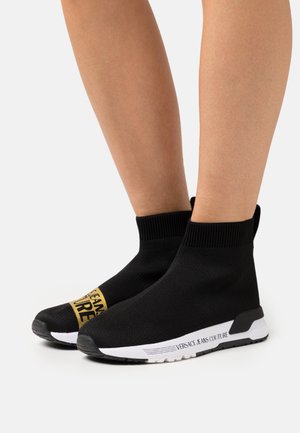 Black knitted ankle boots with a ribbed collar and gold Versace Jeans logo. Features a white sole with black accents.