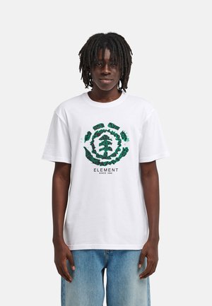 Jeune personne à la peau foncée et aux dreadlocks portant un t-shirt blanc avec un logo graphique d'arbre vert et un jean bleu.