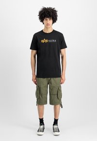 Svart bomull T-skjorte med gul "ALPHA INDUSTRIES" logo, sammen med olivengrønne cargo shorts med flere lommer og snor detaljer.