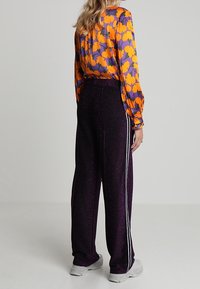 Blusen med långa ärmar har ett mönster av orange och lila blommor; ihop med högmidjade, glittrande lila byxor med vita sidstrimmor.