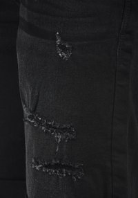 Jean en denim noir avec des détails usés au genou, des bords effilochés et des textures variées dans le tissu.