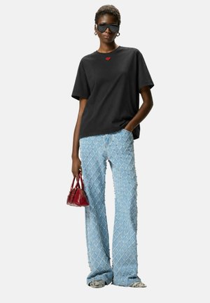 Modella che indossa una T-shirt nera oversize con un piccolo cuore rosso, jeans azzurri con motivo a diamanti, occhiali da sole neri, con una piccola borsa rossa in mano.