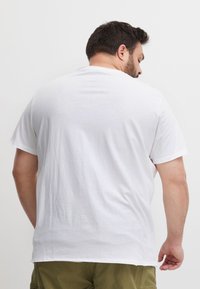 Mand med kort mørkt hår iført en ensfarvet hvid t-shirt og olivengrønne bukser, vendt væk fra en hvid baggrund.