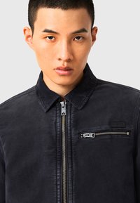 Giacca blu navy con colletto in corduroy, chiusura con zip frontale e tasca sul petto con dettaglio in zip argentato. Realizzata in un materiale liscio e testurizzato.