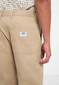 Béžové pracovní kalhoty s obdélníkovým zadním kapsou, na které je modro-bílá etiketa. Odolná látka s hladkou texturou, rovný střih a poutka na pásek.