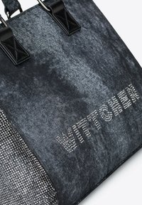 Czarna torba na ramię wykonana z teksturowanej tkaniny z błyszczącymi srebrnymi akcentami, z napisem "WITTCHER" w ozdobnych literach. Czarną skórzaną rączka.