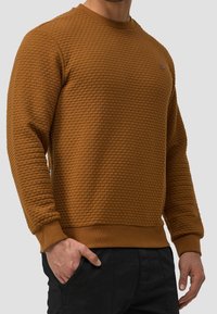 Brązowa bluza z teksturowanym, wypukłym wzorem. Ściągacze na mankietach i dekolcie. Małe logo na piersi. Noszona z czarnymi spodniami.