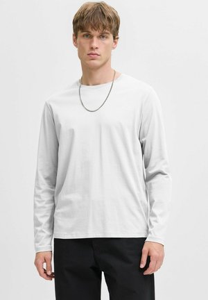 Young man with light brown hair wearing a white long-sleeve shirt, black pants, and a silver chain necklace, standing against a plain background.