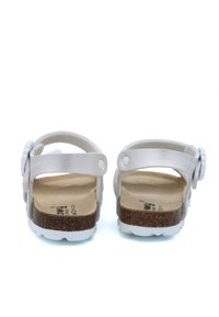 Sandales blanches sans dos avec des brides de cheville réglables, une semelle en liège et une semelle en caoutchouc. Accentées d'une décoration circulaire sur la bride.