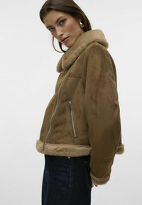 Giacca in fibra scamosciata marrone con collo e polsini in pelliccia. Presenta una tasca con cerniera e un design aderente che crea un contrasto con il denim blu.