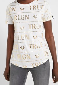 Bílé tričko s krátkým rukávem a zlatým metalickým textem "TRUE RLGN" a motivy podkov, nošené s vybledlými šedými džíny na osobě s tmavou pletí.
