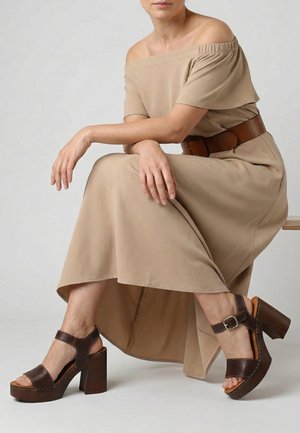 Vrouw zit op een bankje en draagt een beige off-shoulder jurk met een bruine riem en donkerbruine sandalen met hoge hakken en enkelbandjes.