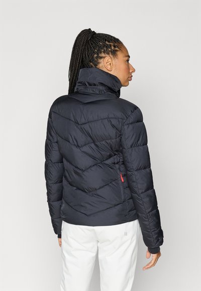 Casaco acolchoado em azul-marinho escuro, com design acolchoado em chevron horizontal, gola alta e fecho de correr vermelho em destaque. Textura lisa e corte ajustado.