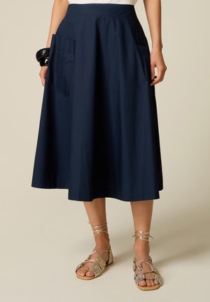 Mujer que lleva una falda midi azul oscuro con grandes bolsillos laterales, sandalias planas doradas con tiras y pulseras negras en una muñeca.
