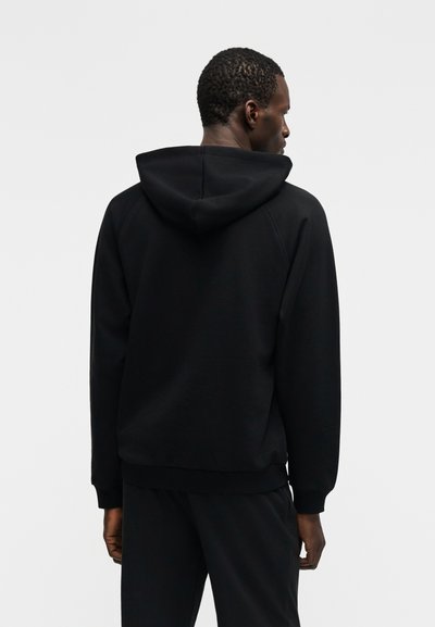 Mann vendt bort med på seg en svart hettegenser og matchende svarte bukser mot en ensfarget lys grå bakgrunn.