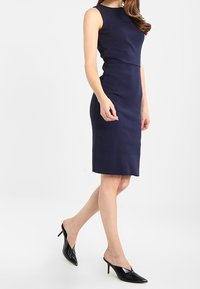 Laivaston sininen hihaton mekko, jossa on tiukka siluetti, sileä kangas, polvenpituinen helma, yhdistettynä mustiin lakattuihin nahkakorkkareihin, joissa on teräväkärkiset varpaat.