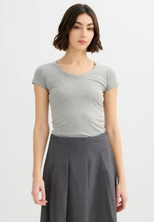 Mujer con cabello corto y oscuro, vistiendo una camiseta de manga corta gris claro con escote en V y una falda plisada de cintura alta en gris oscuro, de pie sobre un fondo blanco.