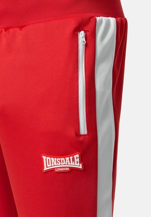 Červené sportovní kalhoty s kapsou na zip, bílým bočním pruhem a výrazným bílým a červeným logem Lonsdale. Hladký povrch látky.
