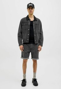 Veste en denim gris foncé avec poches avant, superposée à un débardeur noir. Short gris foncé assorti, chaussures noires et chaussettes grises. Casquette noire.