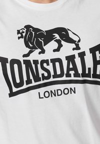 Fehér pamut póló, amelyen egy fekete oroszlán grafika található, valamint vastag betűkkel olvasható a "LONSDALE LONDON" felirat, ívelt díszítéssel a szöveg alatt.