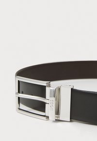 Ceinture en cuir noir avec une boucle en métal argenté comportant deux broches et une gravure de logo. Texture lisse et design minimal.