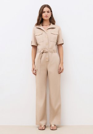 Frau in beigefarbenem Jumpsuit mit Gürtel, kurzen Ärmeln, vorderen Taschen, Knopfdetails und offenem Kragen, stehend vor weißem Hintergrund.