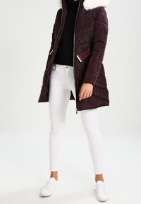 Burgundy vadderad jacka med en foder hoodie, dragkedja fram, sidofickor och en figursydd silhuett, parat med vita skinnyjeans och vita sneakers.