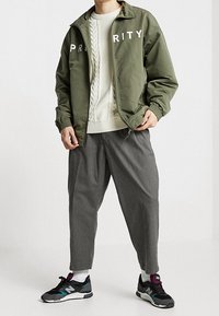 Olijfgroene bomberjack met witte letters die "PRIORITY" spellen, crèmekleurige kabelgebreide trui, grijze tapered broek en zwarte sportieve sneakers.