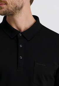 Mann mit schwarzem Poloshirt mit drei Knöpfen und einer Tasche, die das "Pierre Cardin"-Logo zeigt, in der Höhe des Halses und der Brust gekürzt.