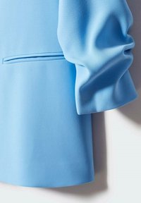 Vaaleansinisestä bleiseristä, jossa on sileä kangas, jämäkkä muotoilu ja rypytettyjä hiharesoreita sekä hillitty sivutasku.