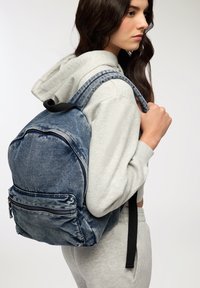 Zaino in denim con lavaggio blu sbiadito, dotato di due spallacci regolabili, una tasca anteriore con zip e forma arrotondata. Finitura in tessuto testurizzato.