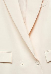 Blazer crema de doble botonadura con solapas anchas y bolsillos de solapa, mostrando detalles de botones en la parte delantera sobre una tela suave.